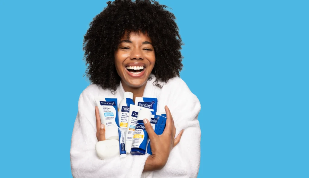 A woman in a white bathrobe smiling and holding all PanOxyl products.