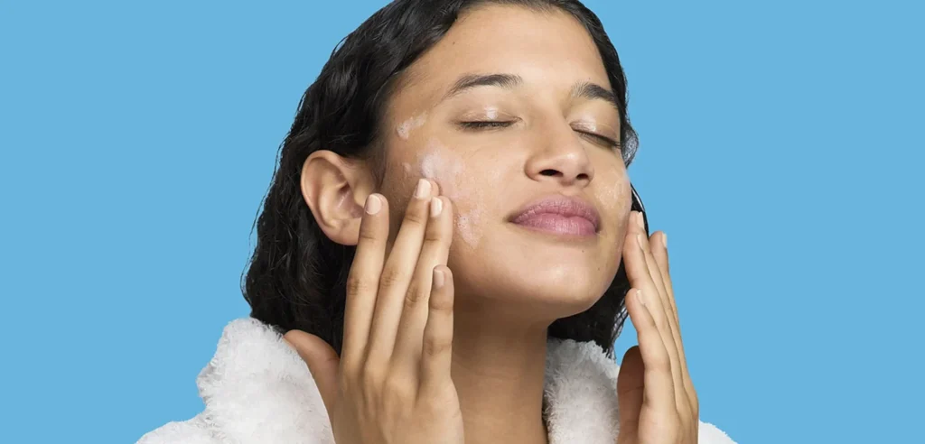 A woman in a white bathrobe applying PanOxyl face wash with her fingertips on a blue background