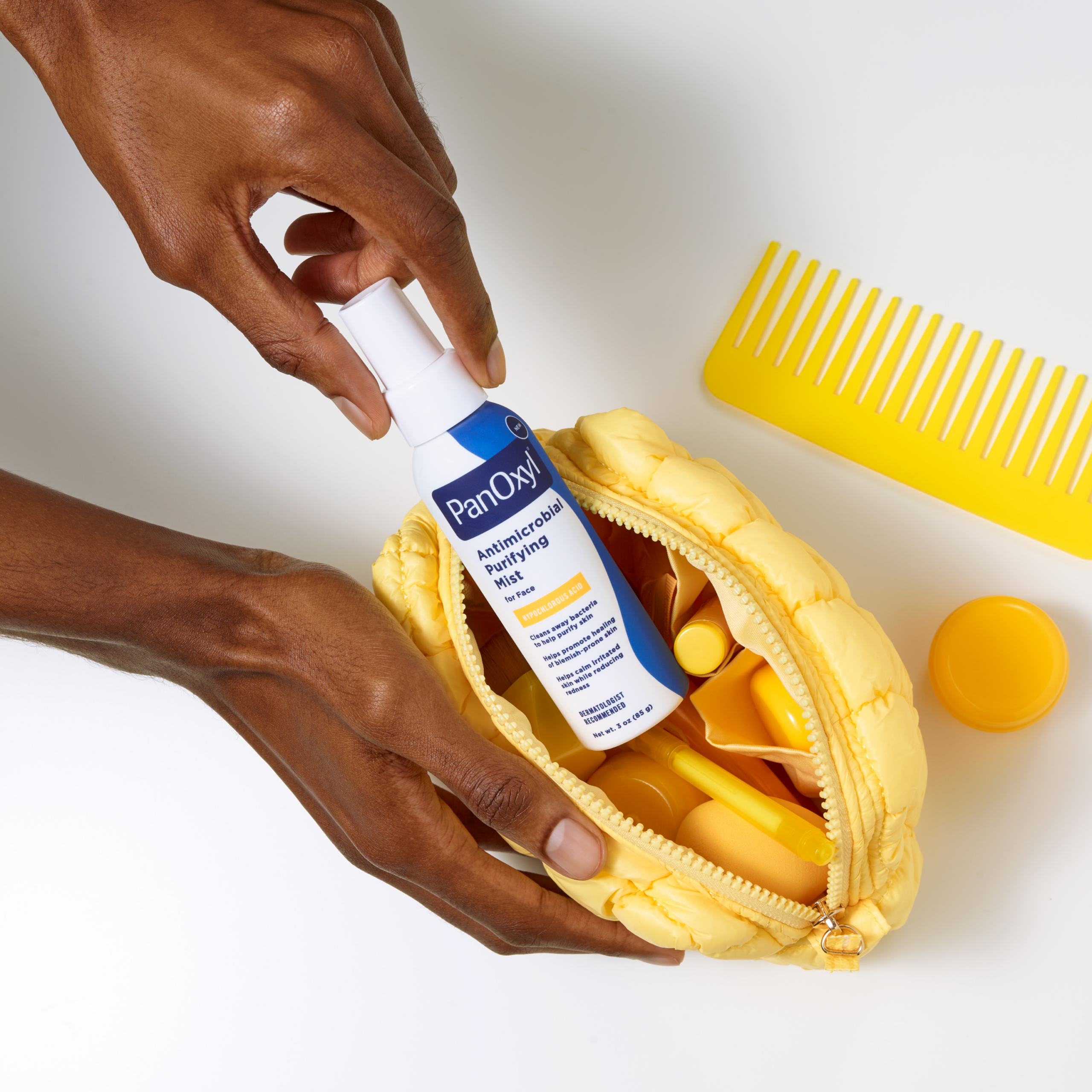 Hand putting PanOxyl Antimicrobial Purifying mist bottle into a yellow makeup bag with other yellow products