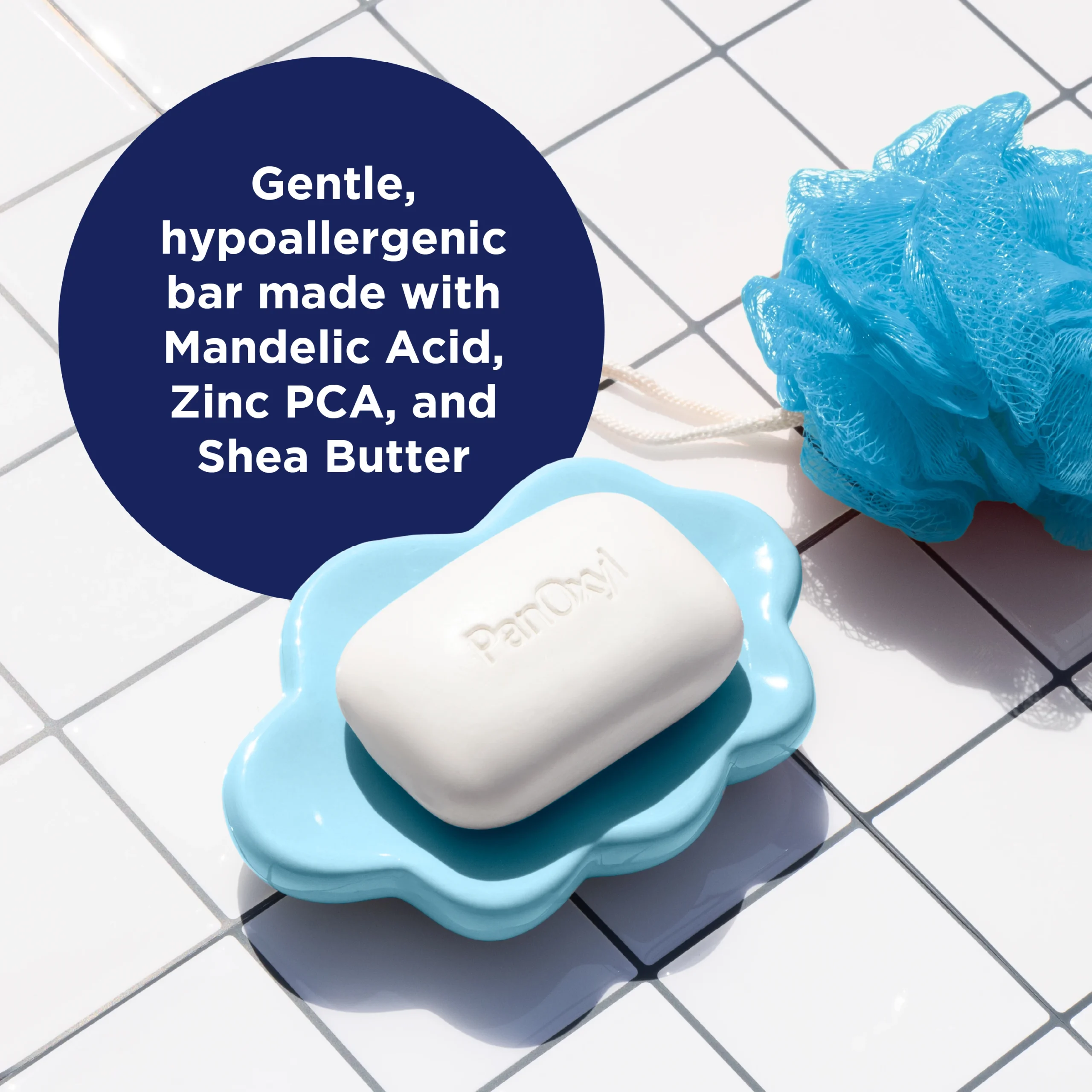 PanOxyl Acne Cleansing bar with 2% Salicylic acid in a blue soap dish next to a blue loofah on a white tile background with text about it's ingredients