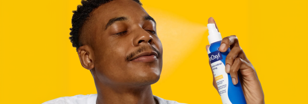 A man spraying PanOxyl Antimicrobial Purifying Mist on his face on a yellow background
